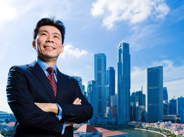 Singapore government minister in a navy suit and red tie, arms folded, photographed against the city skyline