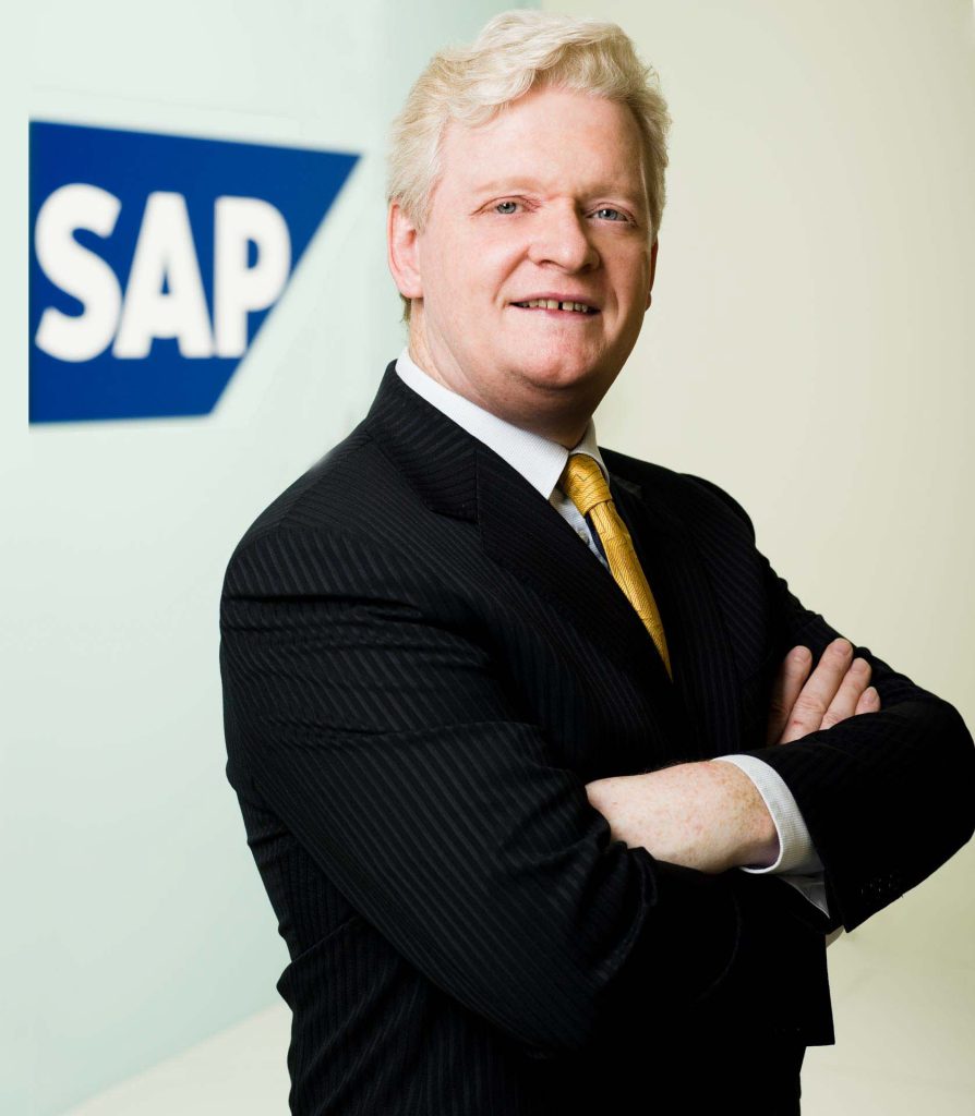 President of SAP in a dark suit and gold tie, arms folded, against the branded SAP blue backdrop