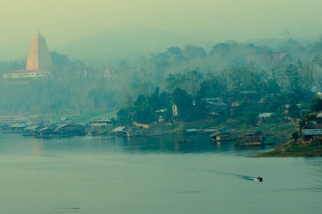 A lone boat crossing a misty lake below a hilltop pagoda and a lakeside village, Thailand — professional travel landscape photography by a Singapore photographer