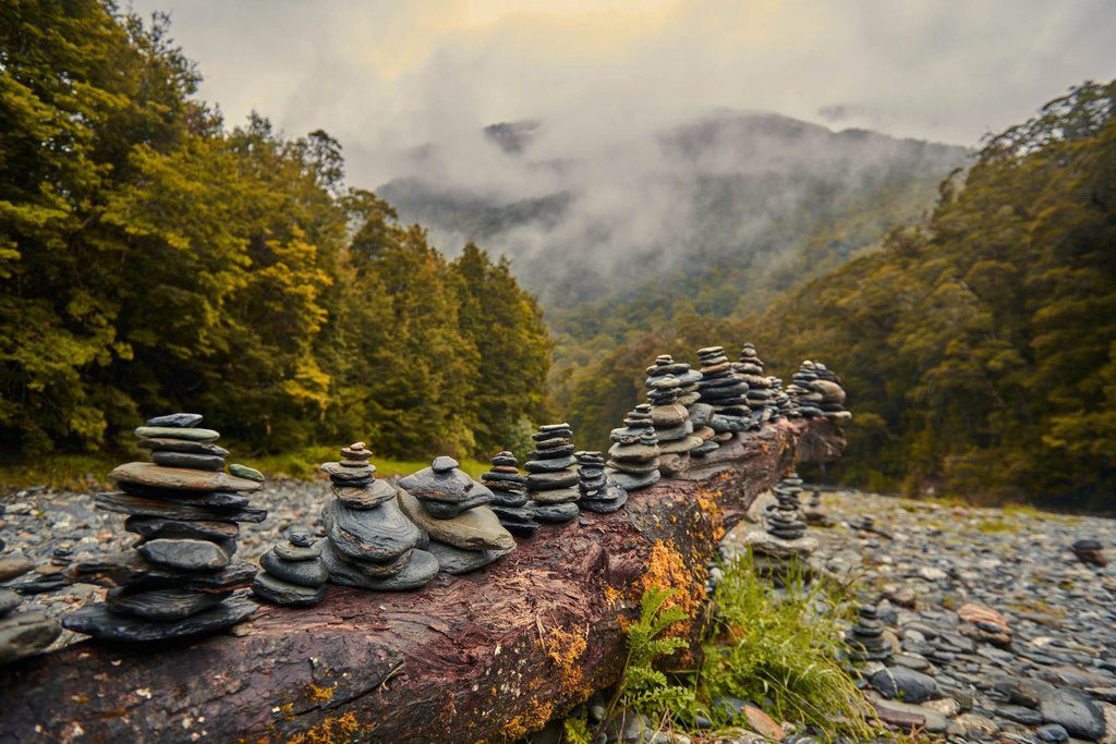Stacked stones on a fallen log beside a stony riverbed, with cloud settling in a forested valley, New Zealand — professional travel landscape photography by a Singapore photographer