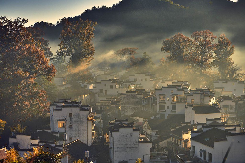 White-walled village houses in morning fog, with sunlight breaking through autumn trees, Wuyuan, China — professional travel landscape photography by a Singapore photographer